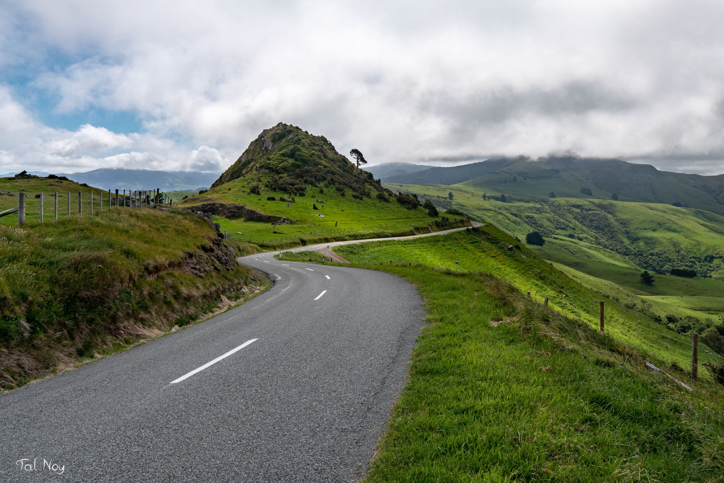 A winding road climbing through lush green hills toward a rocky peak under overcast skies